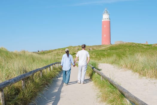 A couple leisurely walking along a coastal path, with a beautiful lighthouse in the background, under a clear sky on Texel Island. man and woman at The iconic red lighthouse of Texel Netherlands
