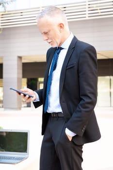 Satisfied with the results, the businessman walks down the street outside the office building, a mature boss holds a phone and coffee in his hands, writes messages and reads news online, using an app.