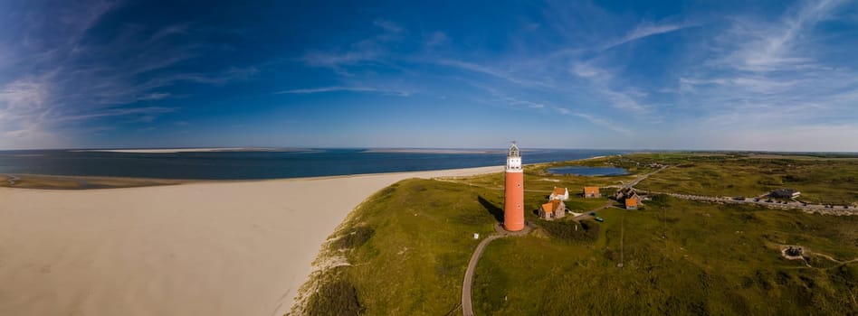 A unique perspective drone view at the lighthouse standing tall on a sandy beach, against a backdrop of the ocean waves crashing. The iconic red lighthouse of Texel Netherlands