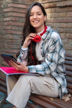 Latin student on campus with headphones surfing the internet with her digital tablet looking at camera. Concept of university life.