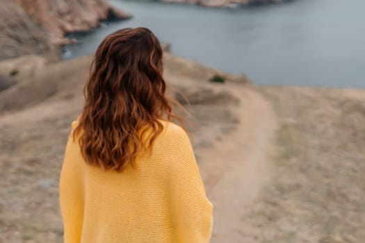 Rear view of a woman with long hair against a background of mountains and sea. Holding a bouquet of yellow tulips in her hands, wearing a yellow sweater.