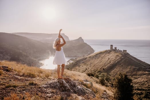 A woman stands on a hill overlooking a body of water. She is wearing a white dress and she is enjoying the view