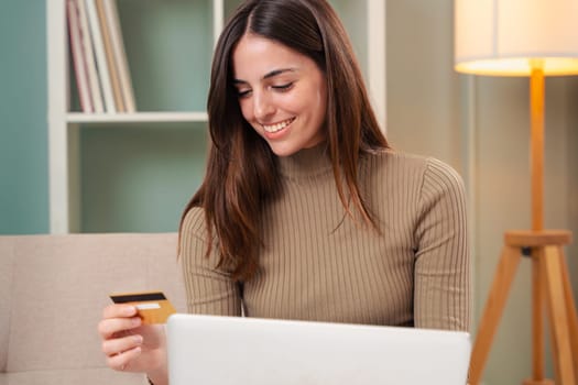 Pretty young caucasian woman shopping online with credit card and laptop while sitting on the couch at home.