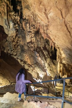 A woman stands on a walkway in a cave, looking up at the ceiling. The cave is filled with stalactites and stalagmites, creating a sense of awe and wonder