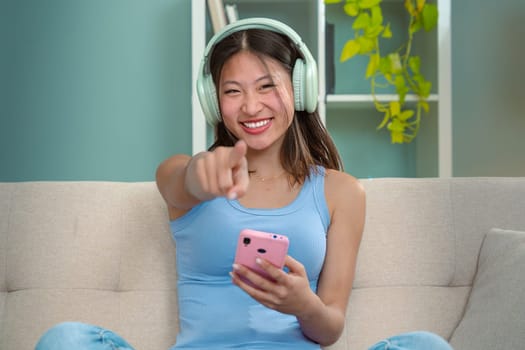 Beautiful woman pointing with thumbs forward, headphones listening to music on sofa happy shopping online with cell phone to make easy and secure remote electronic payments. Inside