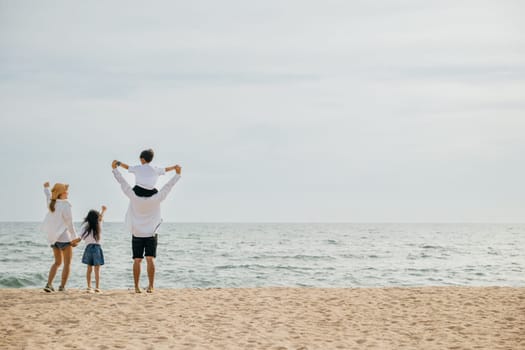 A happy family with raised arms stands on the sea beach at sunset. The father carrying his son on shoulders signifies the carefree joy and togetherness of a perfect beach vacation.