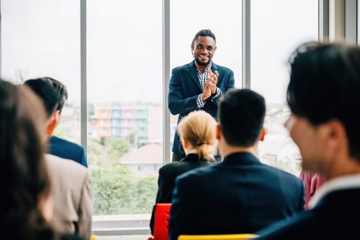 Business professionals from diverse backgrounds meet in an office, with seated team members discussing various topics. Male and female managers stand, guiding the conversation.