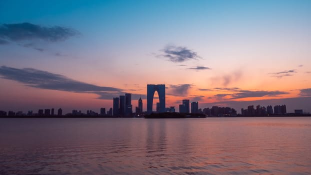 The Gate of the Orient, a distinctive skyscraper in Suzhou, China, is illuminated against the evening sky as it overlooks a calm lake. Surrounding buildings and city lights reflect on the water, creating a picturesque urban scene.