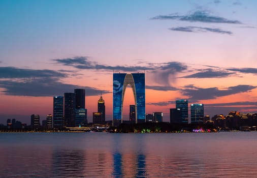The Gate of the Orient, a distinctive skyscraper in Suzhou, China, is illuminated against the evening sky as it overlooks a calm lake. Surrounding buildings and city lights reflect on the water, creating a picturesque urban scene.