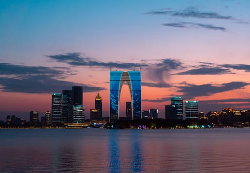 The Gate of the Orient, a distinctive skyscraper in Suzhou, China, is illuminated against the evening sky as it overlooks a calm lake. Surrounding buildings and city lights reflect on the water, creating a picturesque urban scene.