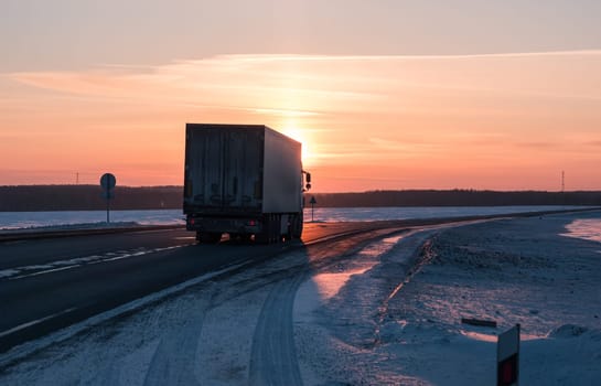 A semi truck cruises down a wintry highway as the sun sets on the horizon, casting a warm glow over the icy road and surrounding landscape.