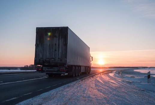 A semi truck cruises down a wintry highway as the sun sets on the horizon, casting a warm glow over the icy road and surrounding landscape.