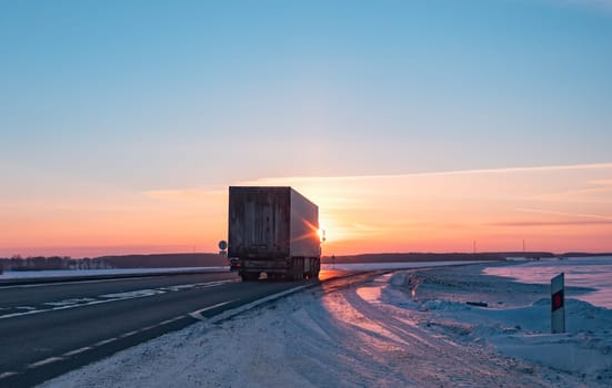 A semi truck cruises down a wintry highway as the sun sets on the horizon, casting a warm glow over the icy road and surrounding landscape.