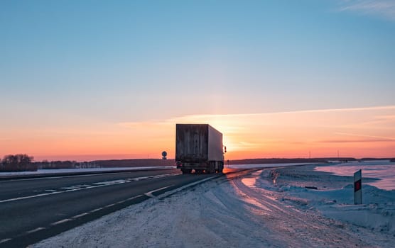 A semi truck cruises down a wintry highway as the sun sets on the horizon, casting a warm glow over the icy road and surrounding landscape.