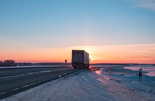 A semi truck cruises down a wintry highway as the sun sets on the horizon, casting a warm glow over the icy road and surrounding landscape.