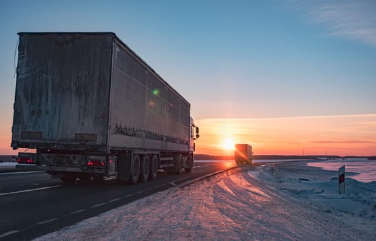 A semi truck cruises down a wintry highway as the sun sets on the horizon, casting a warm glow over the icy road and surrounding landscape.