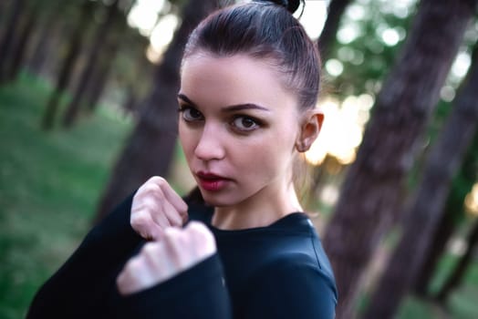 Young woman doing martial arts training in sporty black top holding hands in fighting pose against forest background.