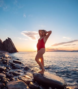 A pregnant woman stands on a rock by the seaside during sunrise.