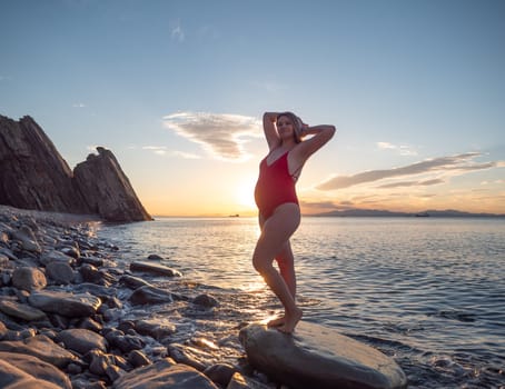 A pregnant woman stands on a rock by the seaside during sunrise.
