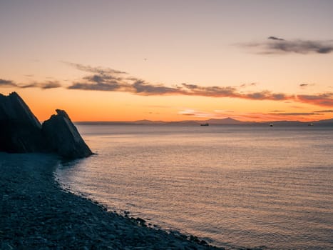 A tranquil sunset casts warm hues over a rocky beach and silhouetted cliffs. The calm ocean waters reflect the changing colors of the sky, creating a serene and picturesque coastal scene.