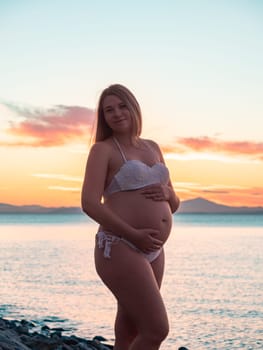 A pregnant woman in a white bikini stands on a rocky beach at sunrise, gently holding her belly. The background features calm water and a distant mountain range.