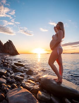 A pregnant woman in a white bikini stands on a rocky beach at sunrise, gently holding her belly. The background features calm water and a distant mountain range.