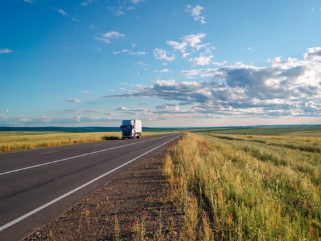 A white truck is seen driving along a long, open road that stretches through a vast countryside under a clear, sunny sky. The surrounding landscape features green fields and distant hills, creating a serene and picturesque setting.
