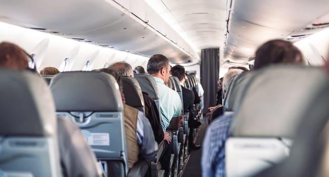 Interior of airplane with passengers on seats and stewardess in uniform walking the aisle, serving people. Commercial economy flight service concept