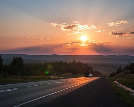 The sun sets in a fiery orange glow over a peaceful road cutting through the countryside.