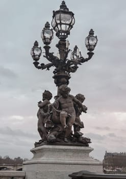 France, Paris - Jan 04, 2024 - a statue of Three naked boys frolicking with fish in hand and at their feet, decorated under four intricately decorated ornate glass lights, and a main central post with a larger glass light on the Alexander III Bridge (Pont Alexandre III) with sky background in Paris. Space for text, Selective focus.