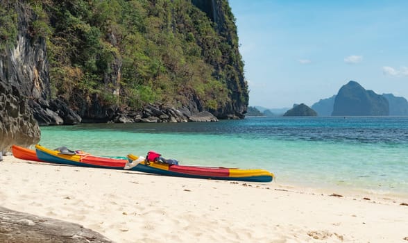 Nestled between towering rocky cliffs and turquoise waters, vibrant kayaks rest on a pristine, sandy beach under a clear, sunny sky.