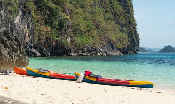 Nestled between towering rocky cliffs and turquoise waters, vibrant kayaks rest on a pristine, sandy beach under a clear, sunny sky.