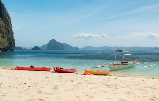 Nestled between towering rocky cliffs and turquoise waters, vibrant kayaks rest on a pristine, sandy beach under a clear, sunny sky.