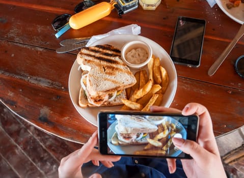 A person is taking a picture of a grilled sandwich and fries at an outdoor cafe.