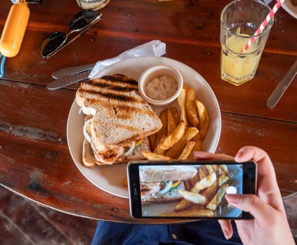 A person is taking a picture of a grilled sandwich and fries at an outdoor cafe.