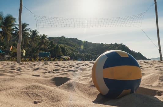 A volleyball rests on the sandy beach beneath a clear, sunny sky. A net is set up, ready for a game. Palm trees and lush green hills are visible in the background.