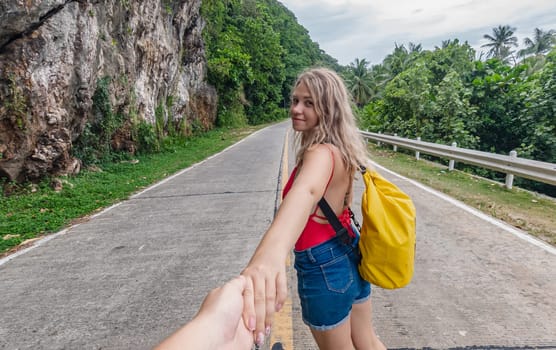 A young woman with long blonde hair, wearing a red top and blue shorts, holds hands with the photographer while walking along a tropical road.