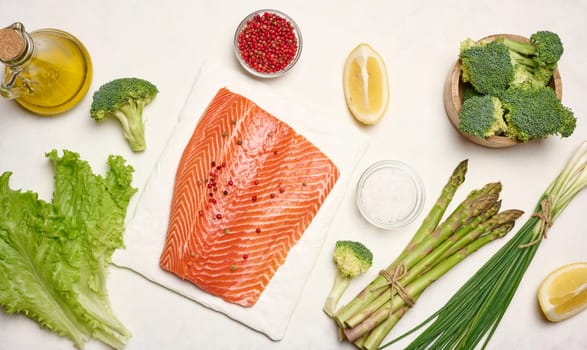 A piece of raw trout fillet on a white board, next to asparagus and broccoli. View from above