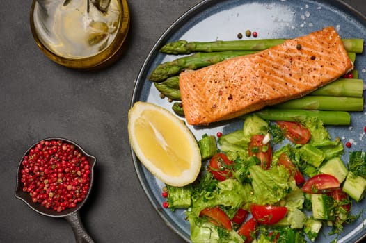 Healthy lunch with grilled salmon with asparagus and fresh tomato and cucumber salad, lemon slice. Food on a blue round plate, top view