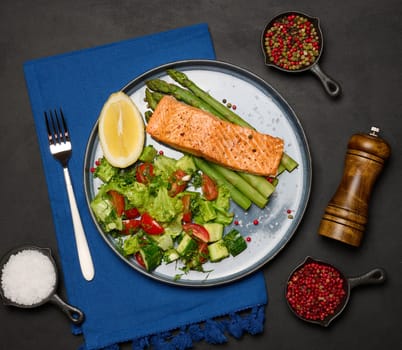 Healthy lunch with grilled salmon with asparagus and fresh tomato and cucumber salad, a lemon slice. Food on a blue round plate, top view
