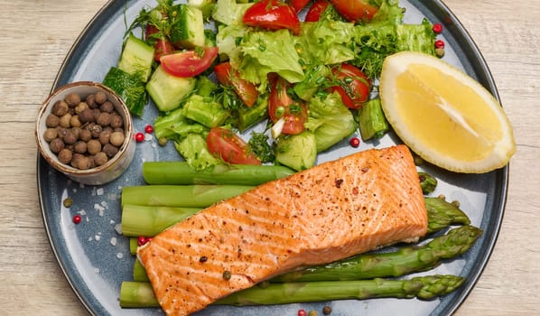 Healthy lunch with grilled salmon with asparagus and fresh tomato and cucumber salad, a row of lemon slice. Food on a blue round plate, top view