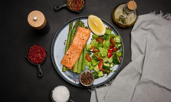 Healthy lunch with grilled trout with asparagus and fresh tomato and cucumber salad, a row of lemon slice. Food on a blue round plate, top view