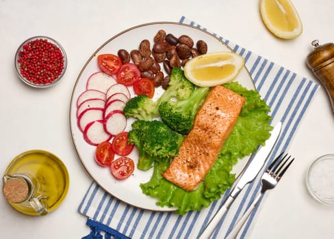 Healthy lunch with grilled trout on green lettuce, next to vegetables, tomatoes, radishes, broccoli and a portion of beans. Accessories include a spice bowl, a bottle of olive oil, a fork and a pepper grinder piece.