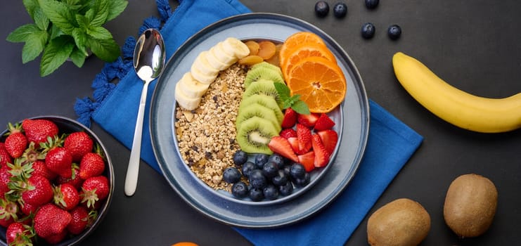 Granola with strawberries, kiwi, banana and blueberries in a round plate on a black table. Healthy and tasty food, top view