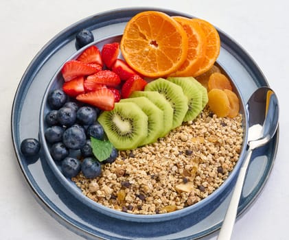 Granola with strawberries, kiwi, banana and blueberries in a round plate on a white table. Healthy and delicious food