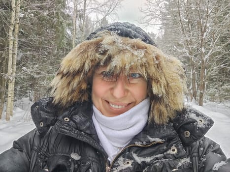 Happy, cheerful middle-aged woman in a warm jacket and a hood with fur taking selfie in winter outdoors. Female Traveler in Alaska, Lapland, Arctic, Siberia