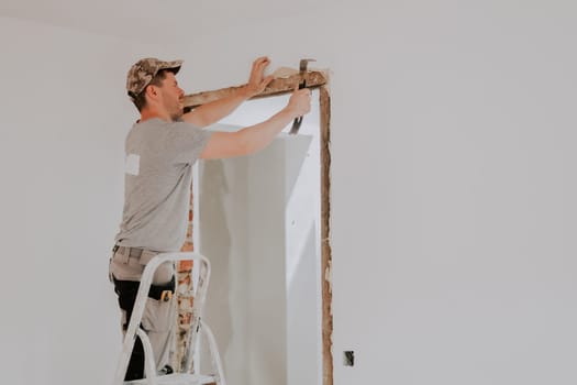 One young Caucasian man in a uniform and cap clears a doorway from old putty using a crowbar while standing on a stepladder in a room with white walls, close-up side view with selective focus. Construction work concept.