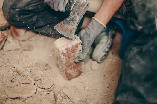 A young caucasian brunette man in black work clothes sits on a small chair on the right and cleans the bricks from dirt, debris and old cement with an ax, top view close-up.Construction work concept.