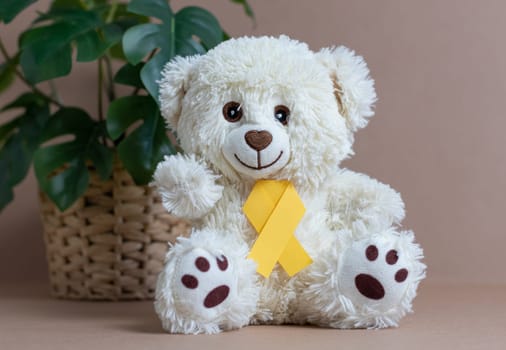 One toy white teddy bear with yellow paper ribbon tie sits near a wicker pot with a flower on a natural nude background, close-up side view. World childhood cancer day concept.