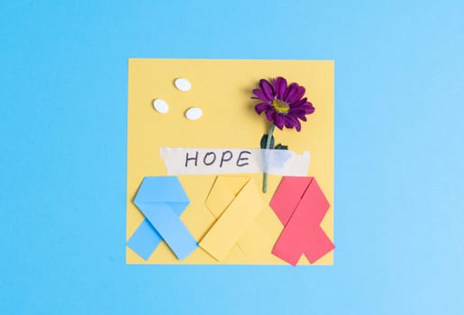 Three multi-colored paper ribbons with a sheet of paper, pills, a gerbera flower and a sticky tape with the inscription hope lie in the center on a blue background with copy space on the sides, flat lay close-up. World Cancer Day concept.